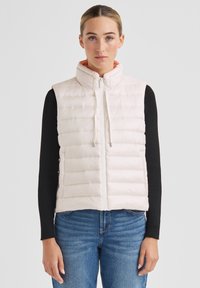 Steppweste in Hellrosa mit horizontalem Steppmuster, hohem Kragen, Frontreißverschluss und Kordelzug-Details. Getragen über einem schwarzen Langarmshirt.