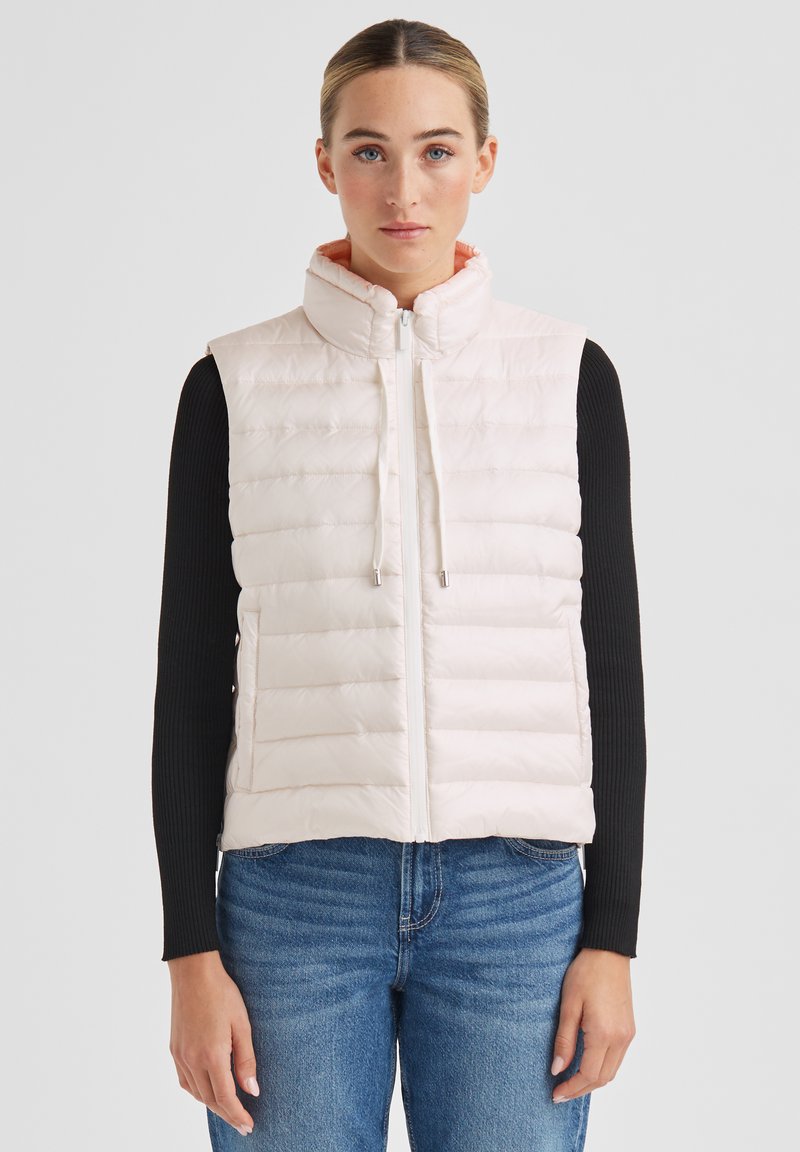 Steppweste in Hellrosa mit horizontalem Steppmuster, hohem Kragen, Frontreißverschluss und Kordelzug-Details. Getragen über einem schwarzen Langarmshirt.