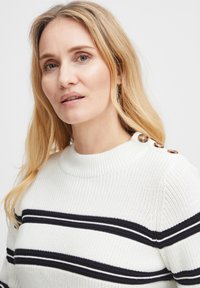 Pull en tricot blanc avec des rayures noires et marines. Il possède un col côtelé et trois boutons décoratifs sur l'épaule gauche.