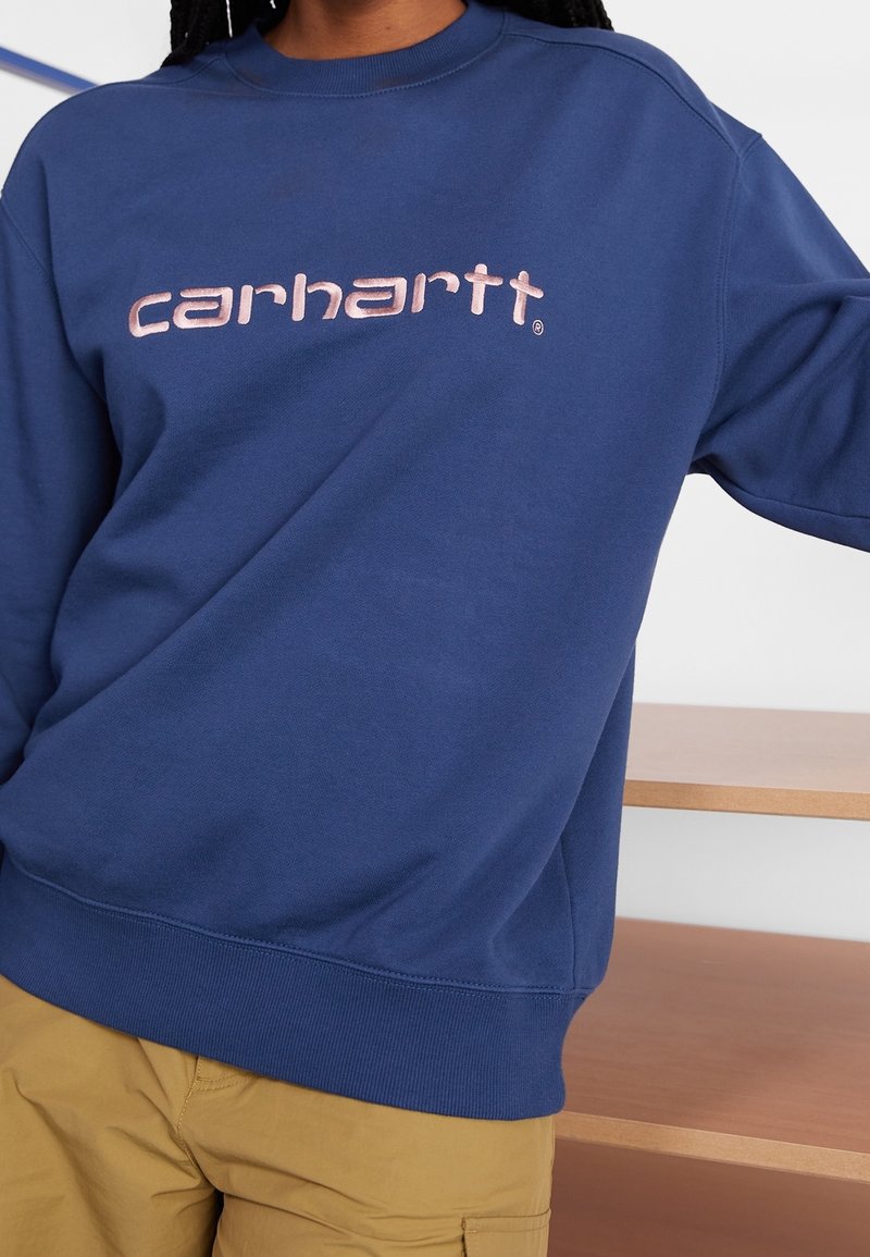 Sudadera azul hecha de una mezcla de algodón, con cuello redondo, puños acanalados y un destacado logo rosa de "carhartt" en el pecho.