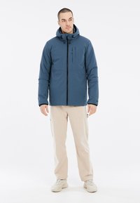 Giacca blu imbottita con cappuccio, zip frontale e tasca sul petto. Indossata con pantaloni beige chiaro e sneakers neutre. Texture liscia.