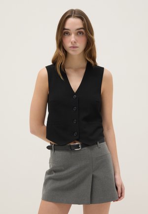 Jonge vrouw met golvend bruin haar die een zwart mouwloos vestje met knopen en een grijze hoge taille rok met riem draagt.