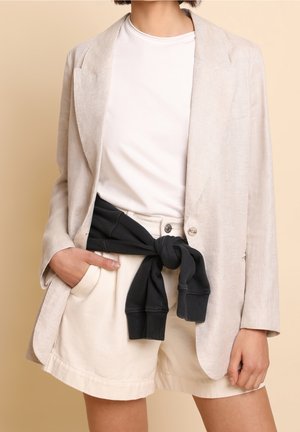 Osoba ubrana w beżową marynarkę na białej koszuli, kremowe szorty z czarną bluzą zawiązaną wokół talii, ręka w kieszeni na beżowym tle.