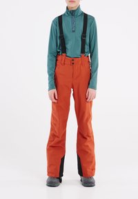 Oranje waterdichte broek met zwarte accenten, voorzien van verstelbare bretels, een ritsvoorkant en een ontspannen pasvorm. Gecombineerd met een teal lange mouwtop.