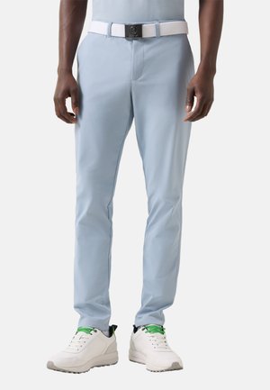 Mand iført lyseblå slim fit bukser, hvidt bælte med sort spænde og hvide sneakers med grønne snørebånd, stående med armene afslappet ned langs siden.