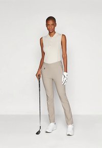 Λεπτή γυναίκα με μπεζ αμάνικο πόλο και ανοιχτό καφέ παντελόνι κρατάει ένα μπαστούνι του γκολφ με το δεξί χέρι, φοράει λευκό γάντι του γκολφ και αθλητικά παπούτσια.