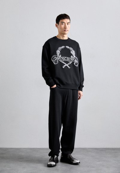 Svart Moschino sweatshirt med vit grafisk logotyp och design detaljer, kombinerat med svarta byxor och mörka skor. Enkel, avslappnad passform.