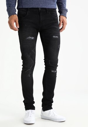 Homme portant un jean slim noir usé, des baskets blanches et une chemise bleue à manches longues, debout les mains relâchées le long du corps.