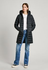 Zwarte gewatteerde lange jas met een capuchon, voorzien van een ritssluiting, zijzakken en een getailleerd ontwerp. Draagt over een wit shirt en denim.