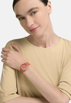 Mujer de piel clara y cabello oscuro lleva una blusa beige y un reloj coral con bisel dorado y marcadores de hora de cristal en su muñeca izquierda.