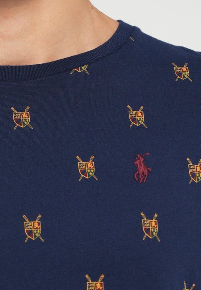 Jūrų mėlynos spalvos medvilnės marškinėliai su mažais išsiuvinėjimais panašiais į skydus ir centriniu raudonu polo logotipu. Klasikinis apvalus kaklas, trumpomis rankovėmis.