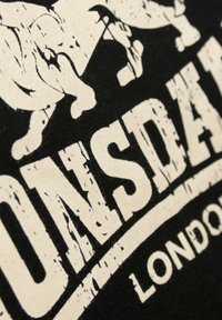 Juodas marškinėlis su balta nusidėvėjusiomis raidėmis "Lonsdale London" ir liūto grafika. Medvilnės medžiaga, apvalus iškirpimas, minkštas tekstūros.