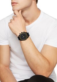 Sort chronografur med en tekstureret sort gummirem, rund urskive, gule accenter og tre sub-ure på en mat sort baggrund.