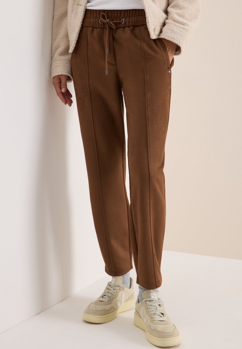Pantalon marron à cordon porté avec des baskets beige et des chaussettes grises, associé à une veste texturée crème sur un fond neutre.