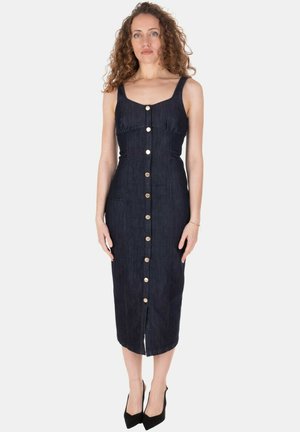 Donna con capelli ricci che indossa un vestito midi aderente in denim scuro senza maniche con bottoni dorati sul davanti e tacchi neri a punta, in piedi su sfondo bianco.