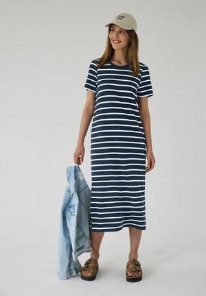 Kvinde iført en navy- og hvidstribet midikjole, beige kasket, brune sandaler, der holder en lyseblå cowboyjakke i venstre hånd.