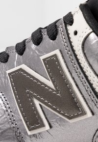 Šedá kovová sportovní bota s texturovaným povrchem, černými tkaničkami a velkým bílým logem "N" na boku. Viditelné zesílené šití.