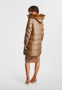 Brązowy długi płaszcz z wypełnieniem i kapturem wyłożonym futrem, z pikowanym wzorem i bocznymi kieszeniami, noszony na spódnicy do kolan i na obcasach.