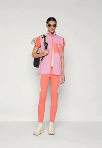 Γυναίκα ντυμένη με κοραλί αθλητικά ρούχα, με ροζ αμάνικο γιλέκο, μπεζ αθλητικά παπούτσια, λευκές κάλτσες, γυαλιά ηλίου και κρατώντας μια σακίδιο.