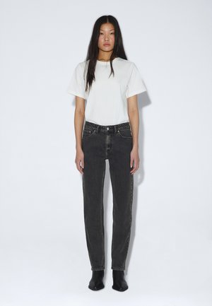 Giovane donna con lunghi capelli neri che indossa una t-shirt bianca, jeans neri a vita alta e stivaletti neri su sfondo bianco.