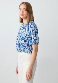 Blusa a manica corta in tessuto azzurro chiaro con motivi astratti blu scuro. Presenta un collo arricciato e una vestibilità ampia. Abbinata a pantaloni bianchi.