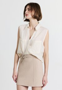 Camicetta senza maniche di colore crema chiaro con righe verticali e due tasche sul petto, abbinata a una minigonna beige con due fibbie per cintura.