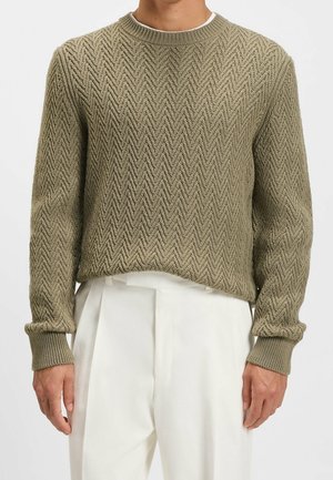 Man in een olijfgroene kabelgebreide trui met chevronpatroon en witte geplooide broek, staand tegen een witte achtergrond.