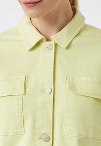 Jasnożółta koszula dżinsowa z ostrym kołnierzykiem, srebrnymi zapięciami na zatrzaski i dwiema kieszeniami na piersi. Tkanina ma gładką fakturę.