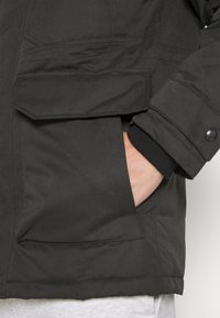 Schwarze, isolierte Jacke mit Reißverschluss und mehreren Fronttaschen; verfügt über elastische Bündchen und eine glatte, strukturierte Außenseite.