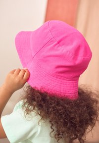 Chapeau bob en coton rose avec un large bord, une texture lisse et des détails cousus, porté par un enfant aux cheveux bouclés.