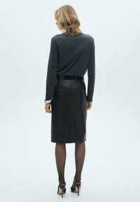 Jupe crayon en cuir noir au design ajusté, associée à un pull sombre. Le modèle porte des collants transparents et des chaussures à talons hauts.