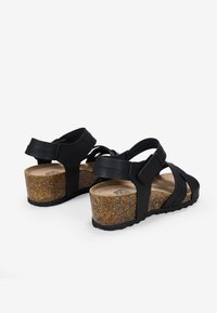 Sandales noires à talon compensé avec des brides entrecroisées, une semelle en liège texturé, et des sangles réglables pour un maintien sécurisé.