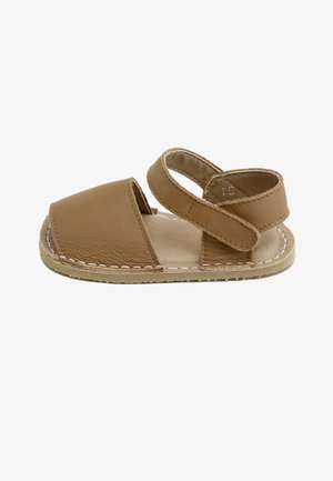 Sandalia marrón para niño pequeño con puntera cerrada, correa ajustable en el tobillo, suela blanda y detalle de costura blanca en el borde de la suela.