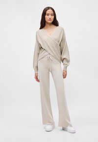 Μπεζ ζακέτα με διαγώνια ραφή και μακριά μανίκια, σε συνδυασμό με παντελόνι φαρδύ που διαθέτει ρυθμιζόμενη ζωνάκι.