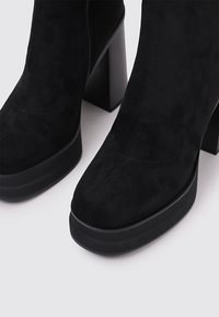 Botines negros de ante con tacón cuadrado sólido, punta redonda y detalle de cremallera en el lateral. Textura suave y diseño minimalista.