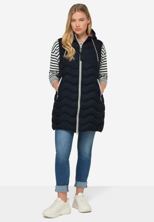 Marine gewatteerde vest met capuchon, voorzijde met ritssluiting en zakken. Draagt over een gestreepte lange mouwen top, gecombineerd met opgerolde jeans en witte sneakers.