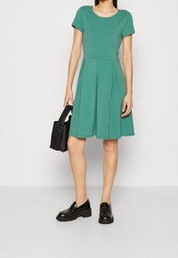 Grön kortärmad klänning med åtsittande midja och utsvängd kjol. Slät tyg, rundad halsringning. Matchas med en svart handväska och kraftiga loafers.