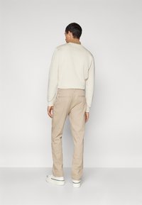Pantaloni di cotone beige e una felpa bianca con un colletto marrone. Sneakers bianche con una suola testurizzata. Sfondo semplice.