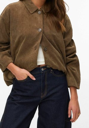 Femme portant une veste en velours côtelé marron boutonnée sur un top court blanc et un jean taille haute bleu foncé, une main dans la poche.