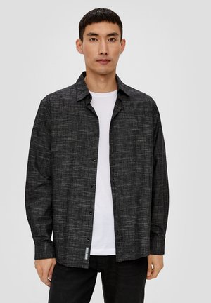 Chemise à boutons noire à motif avec une coupe décontractée, fabriquée en tissu tissé. Elle présente des manches longues et un col classique, portée par-dessus un t-shirt blanc.