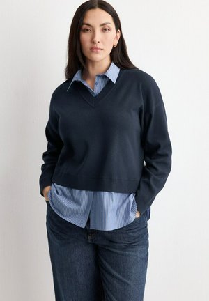 Donna con lunghi capelli scuri in piedi con le mani nelle tasche, indossa un maglione blu navy sopra una camicia a righe blu con bottoni e jeans scuri.