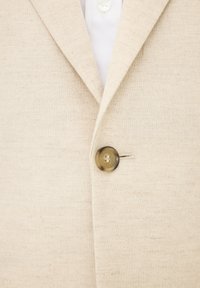 Beige linnen blazer met een gestructureerde afwerking, voorzien van een enkele bruine knoopsluiting en een klassieke reverskraag. Witte blouse eronder.