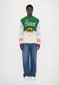 Striktrøje med grønne, blå og pastelfarvede striber, der prydes med "Barrow" og "GOLF" design; parret med brede blå jeans og hvide sneakers.