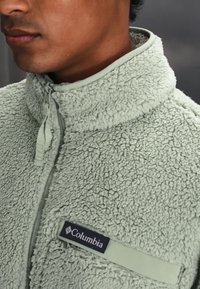 Person trägt eine hellgrüne, strukturierte Fleecejacke mit Reißverschluss und einem Columbia-Logo auf der Brusttasche.