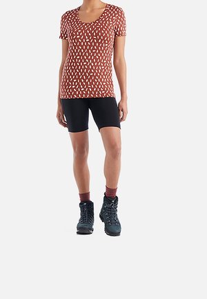 Femme portant une chemise à manches courtes à motifs de couleur rouille, un short noir arrivant aux genoux, des chaussettes bordeaux et des chaussures de randonnée noires.