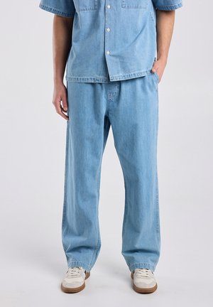 Homme portant un pantalon en denim bleu clair et une chemise en denim à manches courtes assortie avec des baskets blanches, les mains détendues le long du corps sur un fond uni.