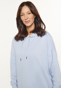 Felpa oversize blu chiaro con maniche lunghe, cappuccio con cordino e dettagli in metallo argentato. Tessuto morbido con una texture liscia.