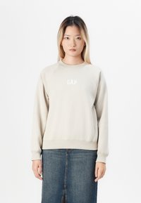 Sudadera de color beige claro con cuello redondo y puños acanalados, con el logo "GAP" en blanco. Combinada con una falda de mezclilla.