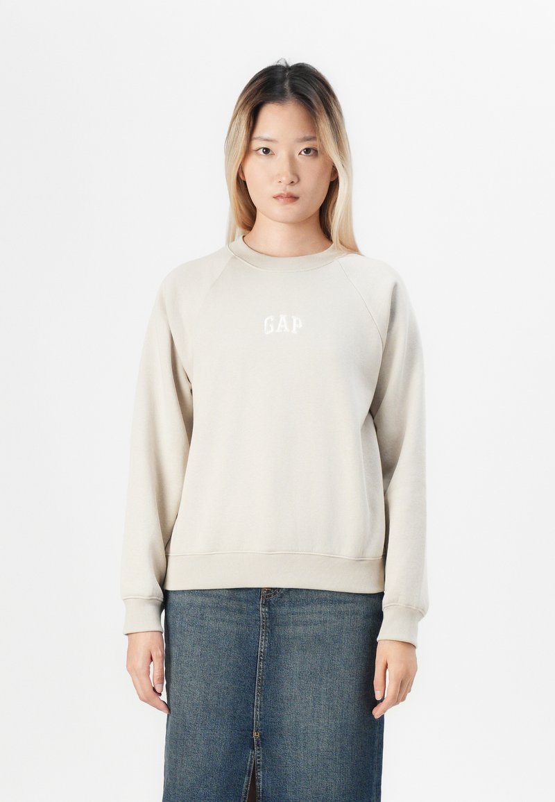 Sudadera de color beige claro con cuello redondo y puños acanalados, con el logo "GAP" en blanco. Combinada con una falda de mezclilla.