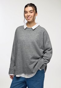 Maglione grigio con una texture morbida, polsini a coste e una vestibilità rilassata, indossato sopra una camicia bianca con colletto, abbinato a jeans in denim blu.